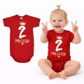 dziewczynka w czerwonym body dziecięcym z białą cyferką dwa. Na paluszkach pokazuje ile ma latek